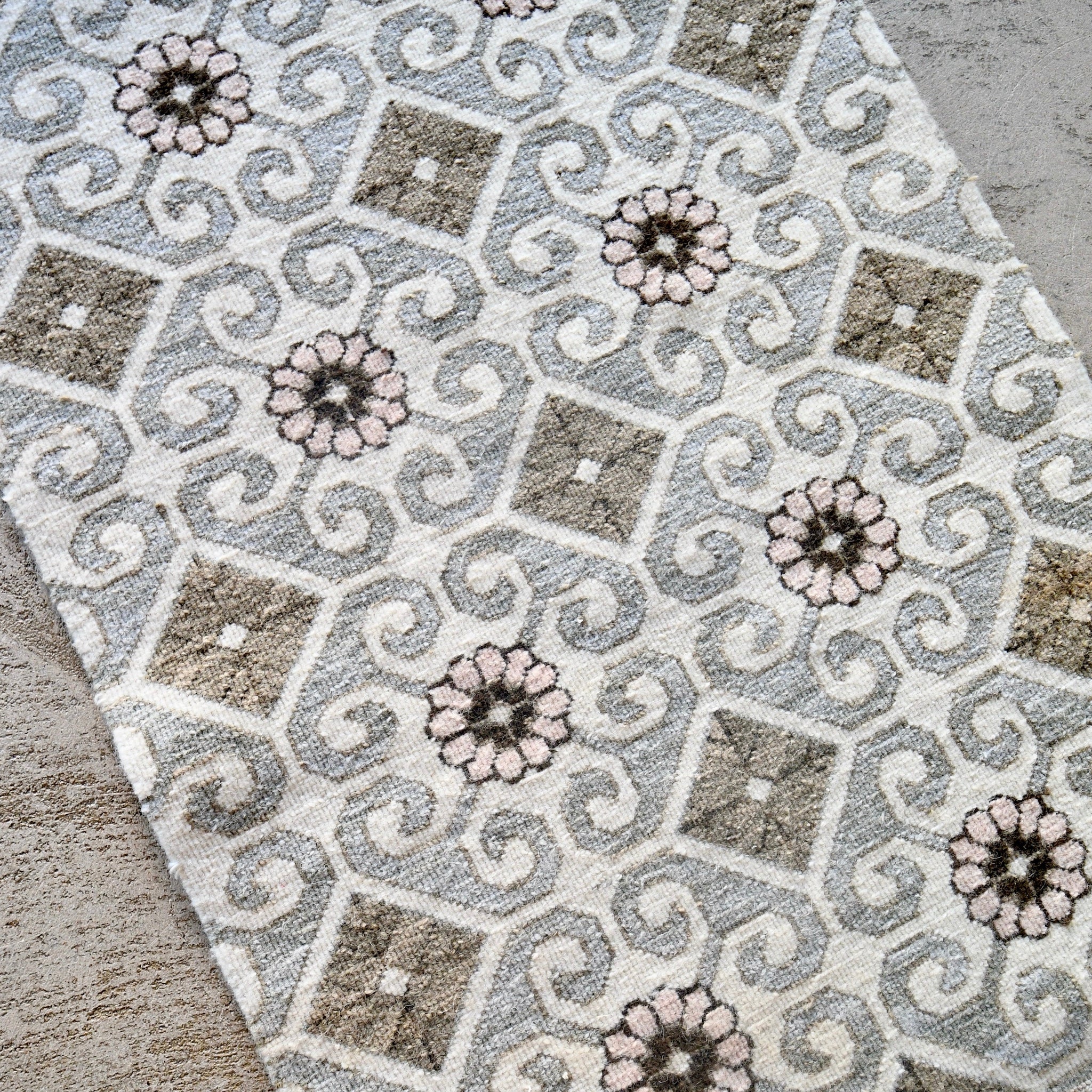 A close-up of a woven textile with an intricate geometric and floral pattern in gray, beige, white, and muted pink, laid diagonally across a neutral surface.
