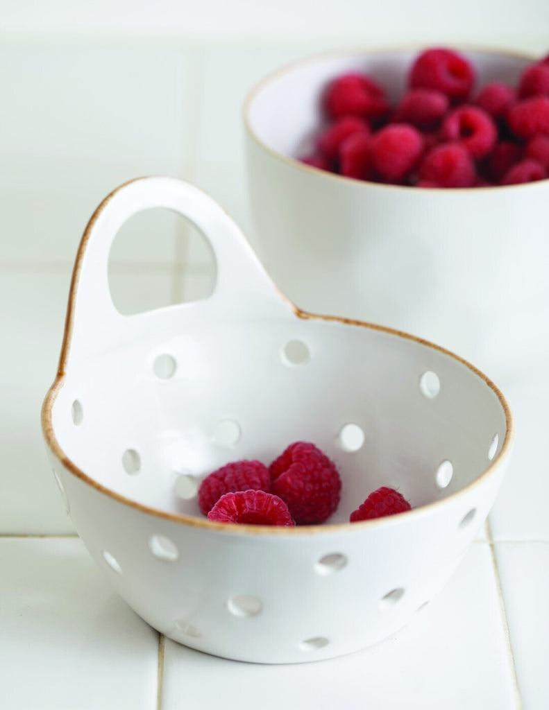 White stoneware colander with raspberries and a BerryHome bowl with additional raspberries in the background.