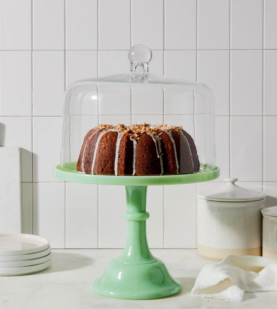 A bundt cake with glaze on a green cake stand, showcased under the Elegant 12" Hand-Blown Glass Cake Dome by Mosser Glass, set against a tiled kitchen backdrop, elevating the dining experience.