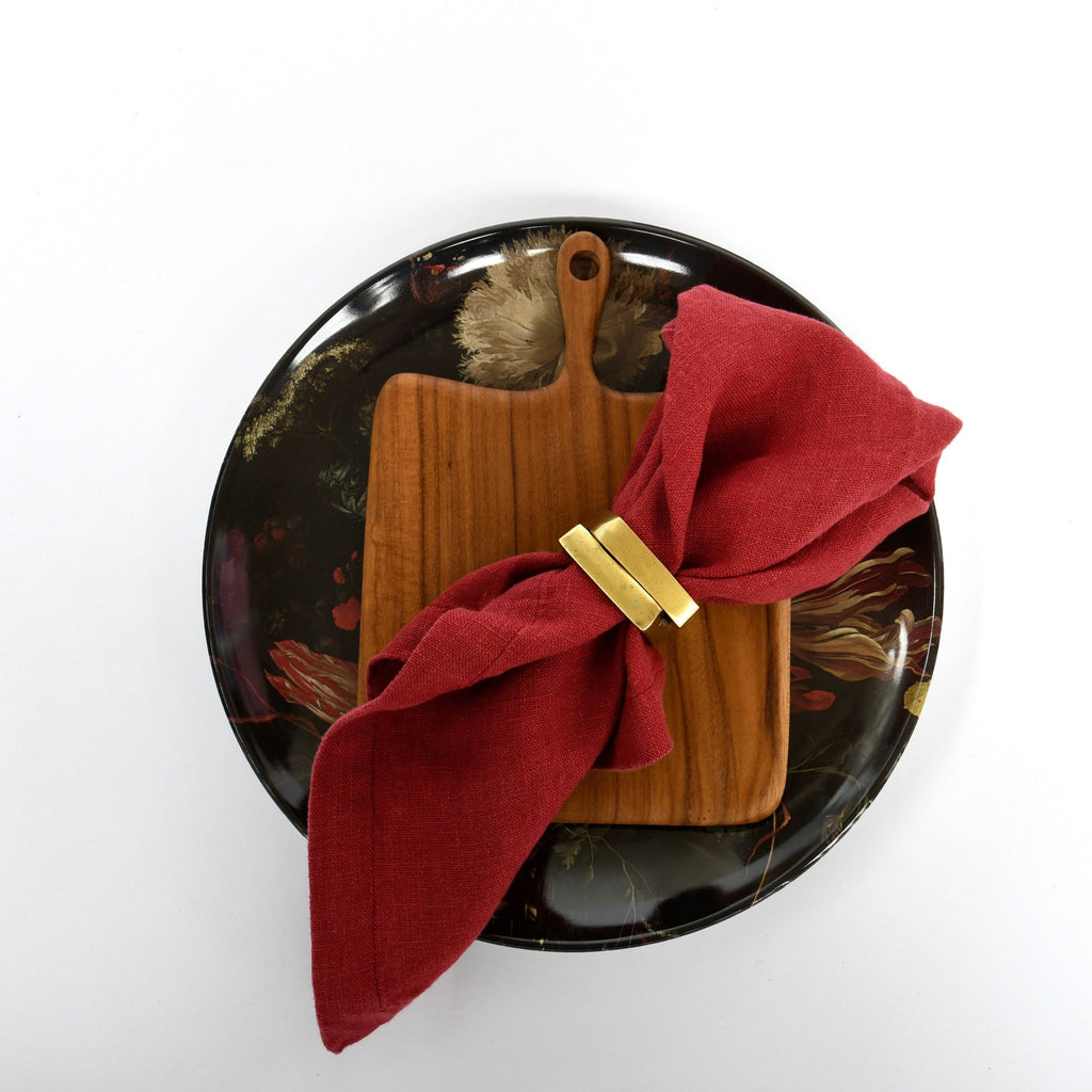 A fancy napkin arranged through Bloomingville Brass Tone Square Napkin Rings - Set of 4, resting on a wooden board atop a decorative plate.