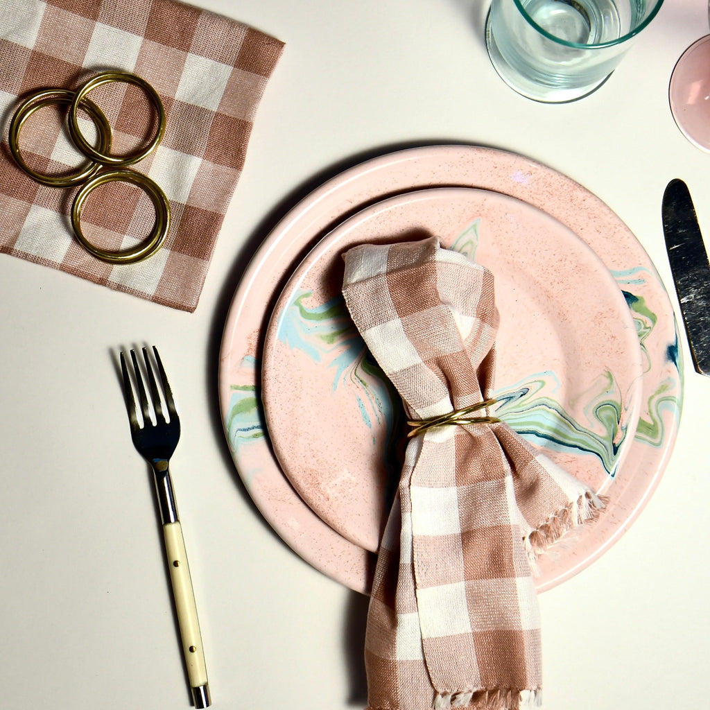 A neatly arranged dining setup with a pink marbled plate, cutlery, and a glass on a light surface, accented with Bloomingville brass napkin rings on leather tie holding a checkered napkin.