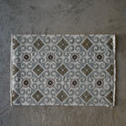 A rectangular woven rug with a geometric tile-inspired pattern in gray, beige, and brown, laid flat on a textured gray concrete surface.
