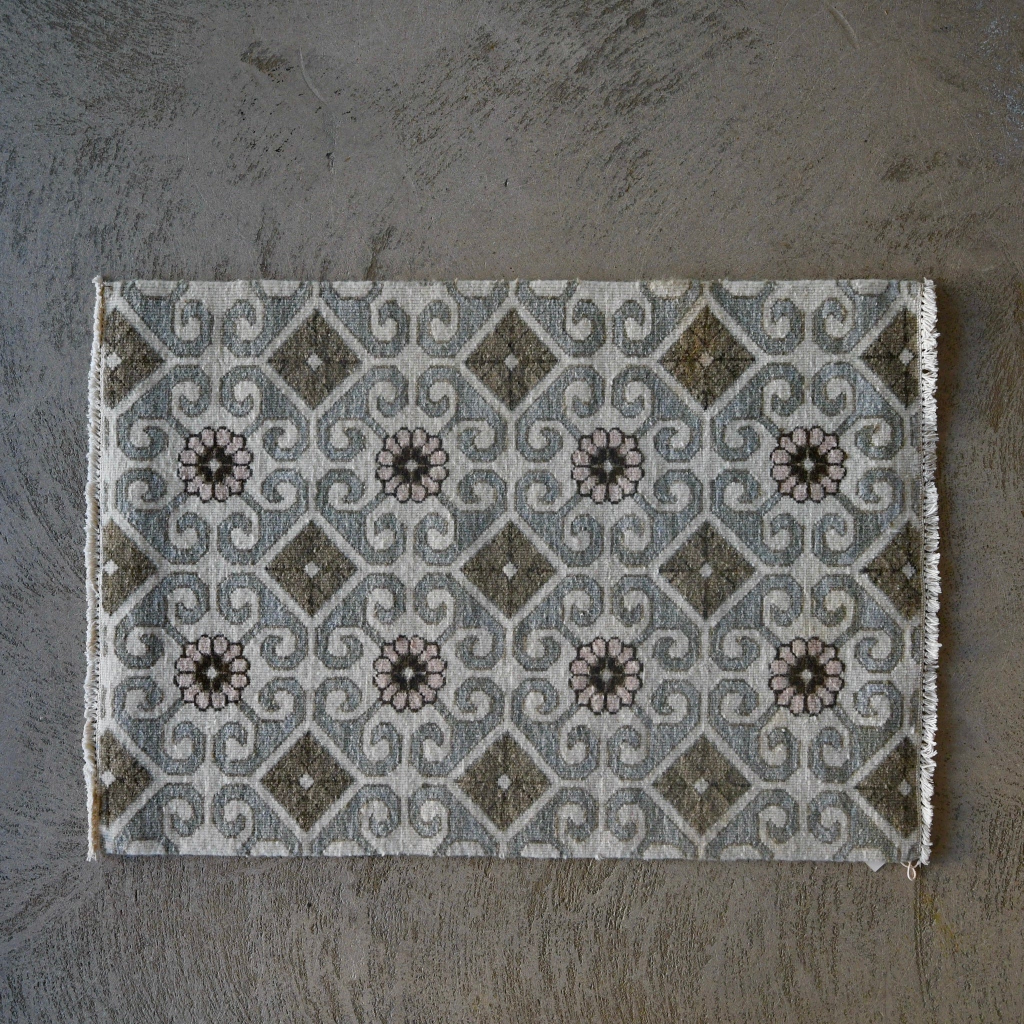 A rectangular woven rug with a geometric tile-inspired pattern in gray, beige, and brown, laid flat on a textured gray concrete surface.