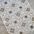 A close-up of a woven textile with an intricate geometric and floral pattern in gray, beige, white, and muted pink, laid diagonally across a neutral surface.