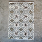 A rectangular area rug with a geometric pattern of gray and brown diamonds, swirls, and floral motifs on a cream background, laid flat on a textured gray surface.