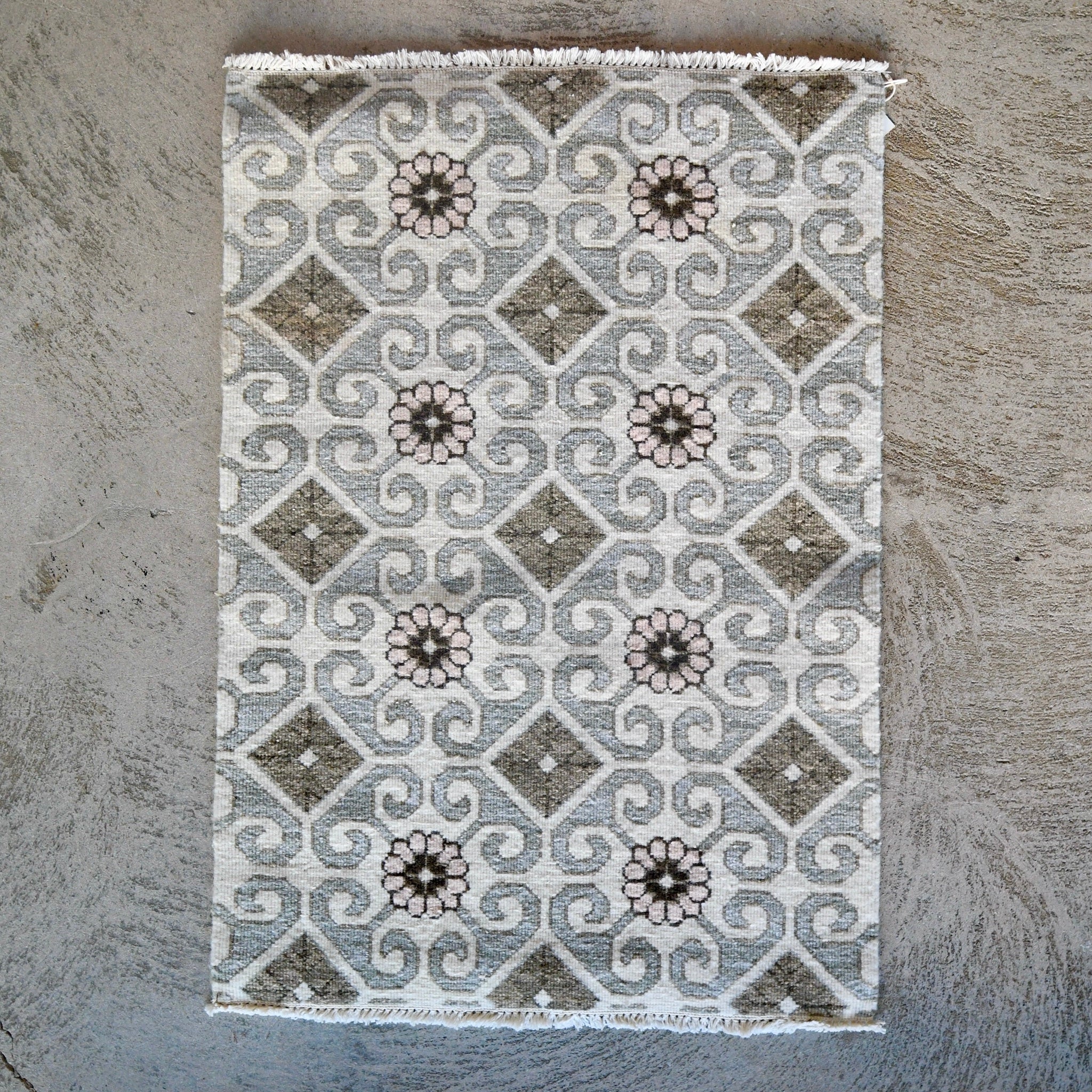 A rectangular area rug with a geometric pattern of gray and brown diamonds, swirls, and floral motifs on a cream background, laid flat on a textured gray surface.