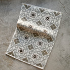 A rectangular woven rug with a geometric and floral pattern in gray, beige, and white, shown laid flat on a textured concrete surface.