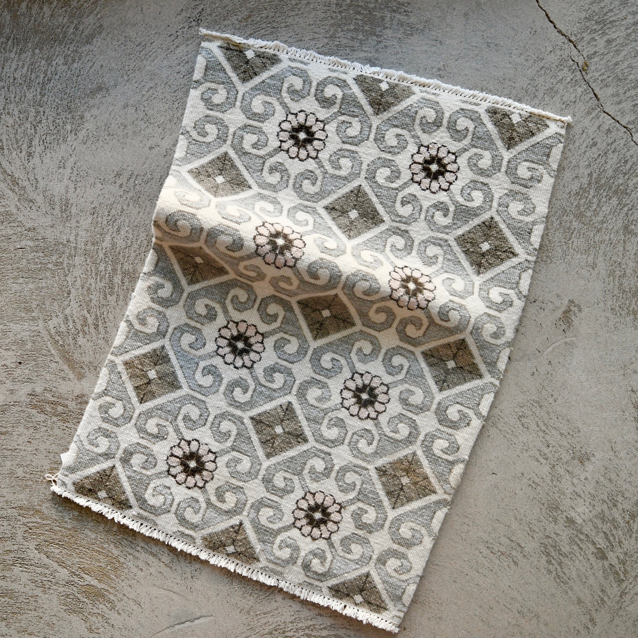 A rectangular woven rug with a geometric and floral pattern in gray, beige, and white, shown laid flat on a textured concrete surface.