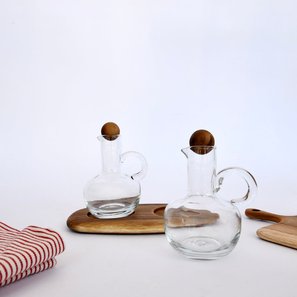 Two handmade in Thailand glass pitchers, an BeHome Acacia Wood Oil & Vinegar Cruet Set with Modern Tray on a table, accompanied by a striped towel and wooden utensils.