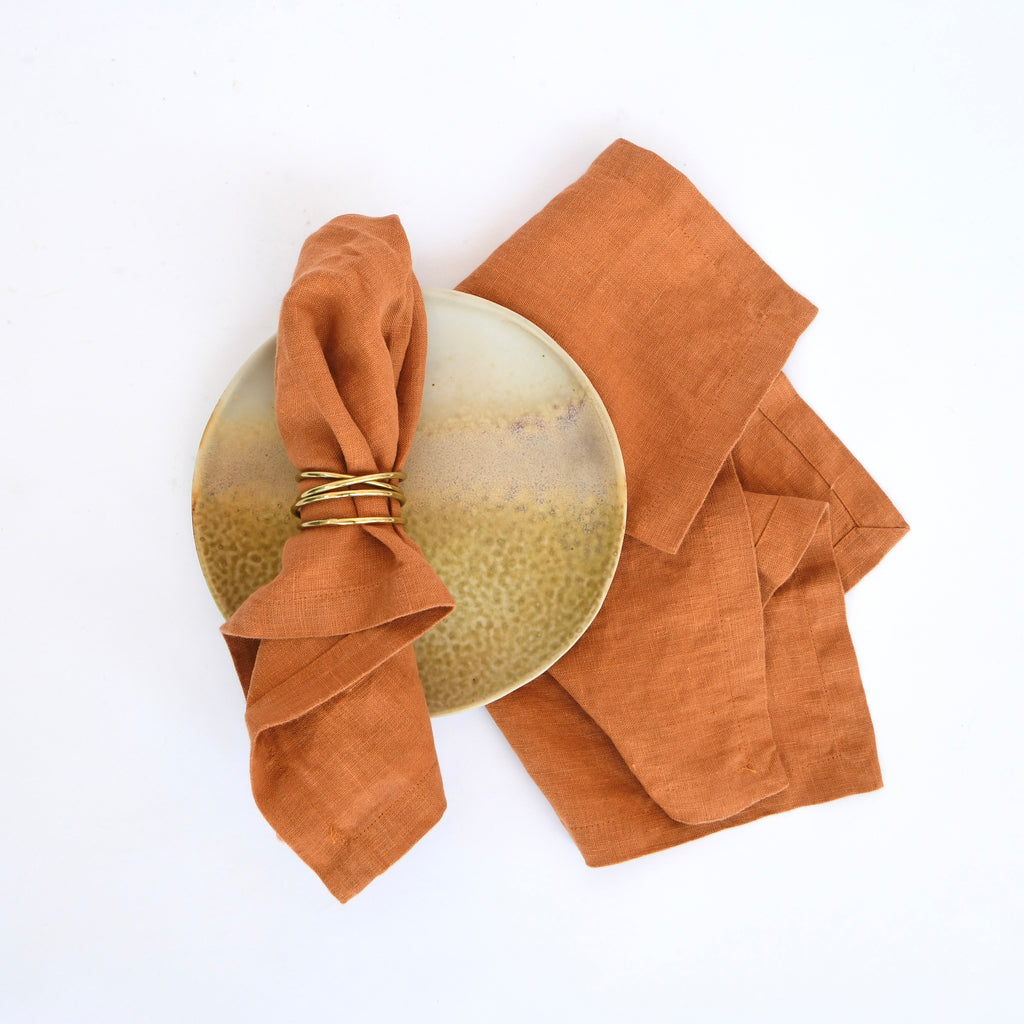 A ceramic plate with a napkin ring and a folded Linen Tales Spiced Orange Linen Napkin on a white background.